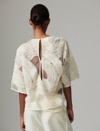 Διάφανο λευκό τοπ με υφή κυματιστού υφάσματος, με άνοιγμα στο πίσω μέρος και κοντά μανίκια, φορεμένο πάνω από απλό λευκό κάτω μέρος.