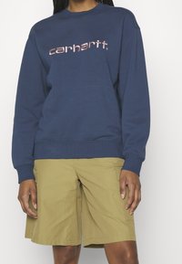 Sudadera azul marino de mezcla de algodón con puños y dobladillo acanalados, que presenta un logo rosa de Carhartt. Combinada con pantalones cortos de cargo beige claro.