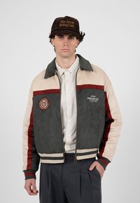 Homme portant une veste de course à blocs de couleur, une chemise rayée, un pantalon foncé et une casquette marron avec l'inscription "L'art d'être en retard".