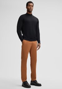 Schwarzes langärmeliges Rollkragenoberteil, braune gerade geschnittene Hose und schwarze Schnürschuhe. Hat ein eng anliegendes Design und eine glatte Textur.