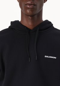 Nærbilde av en person som har på seg en svart Salomon-hettegenser med snøring, med fokus på brystet og den nedre delen av ansiktet.