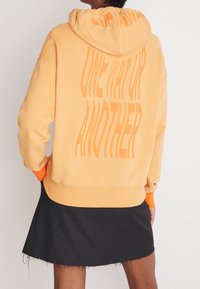 Persona che indossa un hoodie pesca con la scritta "ONE WAY OR ANOTHER" sulla schiena e una gonna nera con l'orlo sfrangiato, di spalle rispetto alla fotocamera.