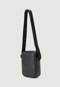 Černá kožená crossbody taška s obdélníkovým tvarem, hladkým povrchem a nastavitelným látkovým popruhem. Minimalistický design, žádné viditelné kovové prvky.