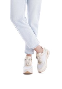 Zapatillas deportivas blancas con acentos en beige claro, paneles de tela texturizada y un logo dorado. Combinadas con jeans de mezclilla azul claro.