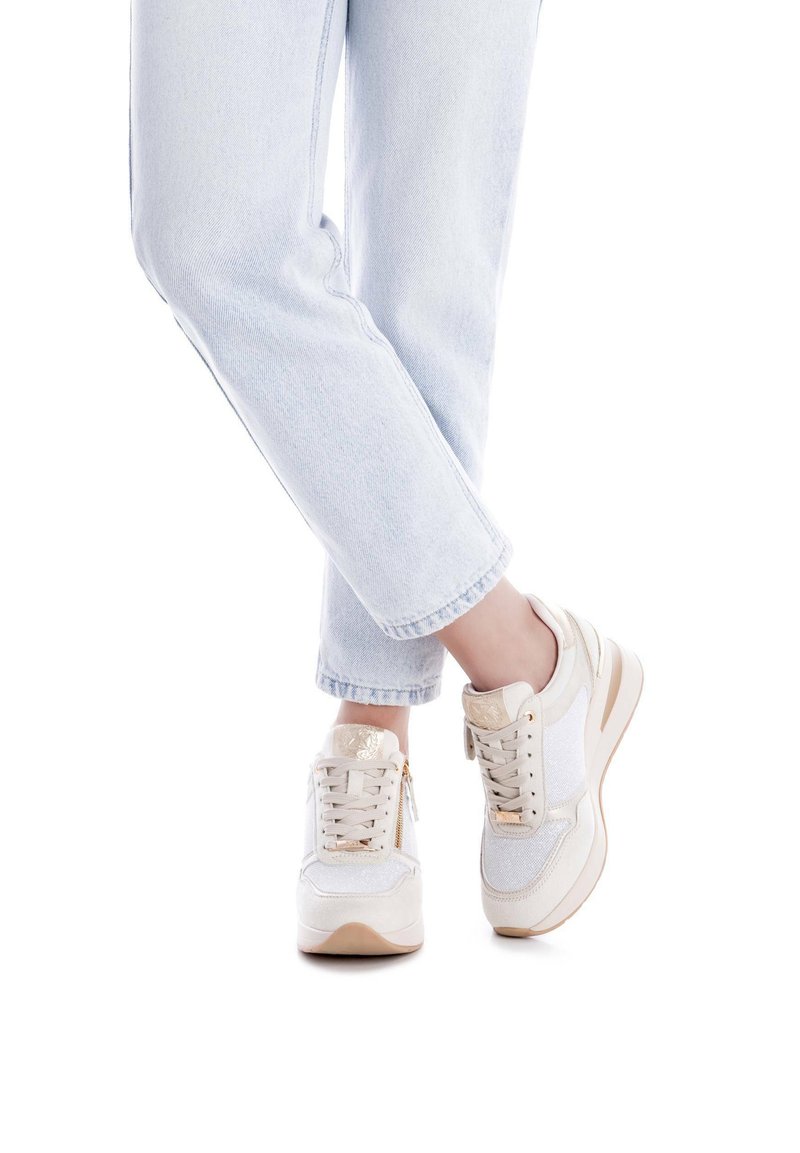 Zapatillas deportivas blancas con acentos en beige claro, paneles de tela texturizada y un logo dorado. Combinadas con jeans de mezclilla azul claro.
