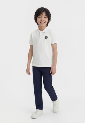 Băiat zâmbitor care merge, purtând un tricou polo alb cu un mic logo negru, pantaloni bleumarin închiși și adidași albi pe fundal alb simplu.