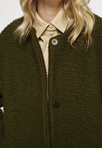 Cardigan verde con trama testurizzata e finitura morbida. Presenta due bottoni neri e una scollatura profonda, indossato sopra una camicia beige con colletto.