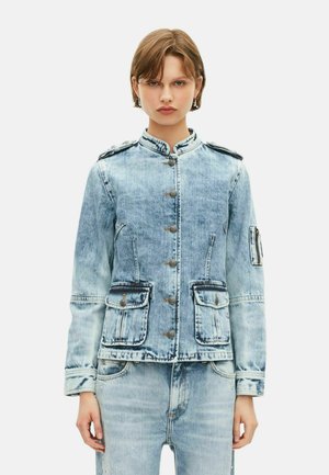 Jongere die een lichtblauwe acid-wash spijkerjack met knoopsluiting, klepzakken en bijpassende acid-wash jeans draagt, tegen een effen achtergrond.