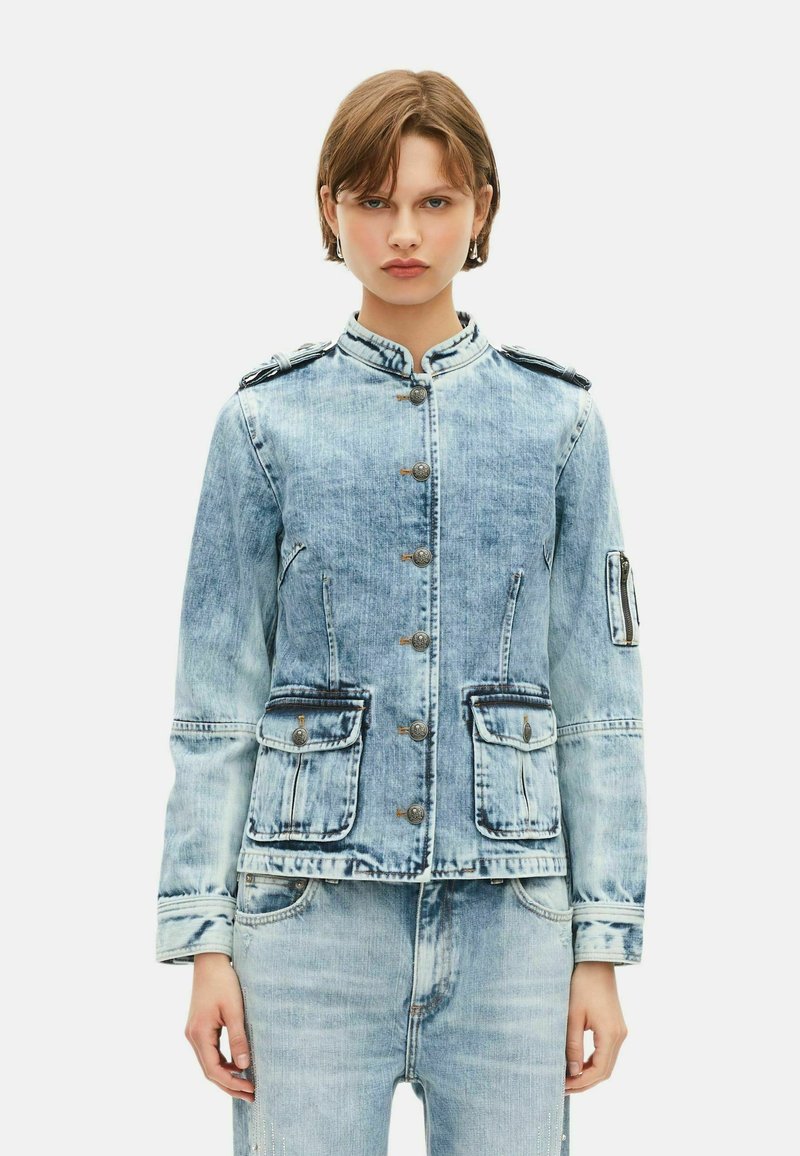 Jeune personne portant une veste en denim bleu clair délavé avec des boutons à l'avant, des poches à rabat et un jean délavé assorti, le tout sur un fond uni.