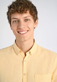 Lichtgele button-up shirt met een zak, met een gestructureerde stof en ronde kraag. Model heeft lichtbruin krullend haar en blauwe ogen.