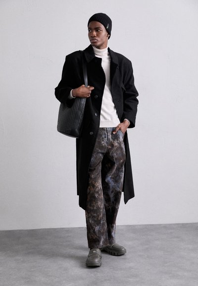 Casaco preto, suéter creme, calças largas estampadas em tons de castanho e azul, gorro preto de malha, bolsa de textura preta, sapatos cinzentos.