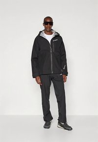 Czarna wodoodporna kurtka z szarym wnętrzem, pełny zamek błyskawiczny, kieszeń na piersi na zamek oraz dopasowane czarne spodnie. Noszona z okularami przeciwsłonecznymi i butami do wędrówek.