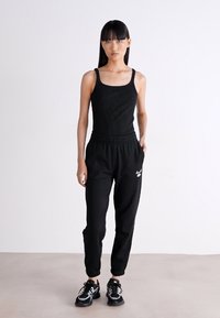 Juodas sportinis kombinezonas su tekstūruotu, prigludusiu viršumi ir elastinga juosmens juosta. Yra juodos spalvos jogger stiliaus kelnės ir baltos logotipo detalės.