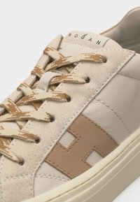 Gros plan d'une basket décontractée beige et marron clair avec des lacets tissés, des détails en daim et un grand "H" cousu sur le côté.