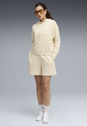 Mujer de pie vestida con sudadera con capucha beige y pantalones cortos, zapatillas blancas, calcetines blancos y gafas de sol con lentes amarillas contra un fondo gris.