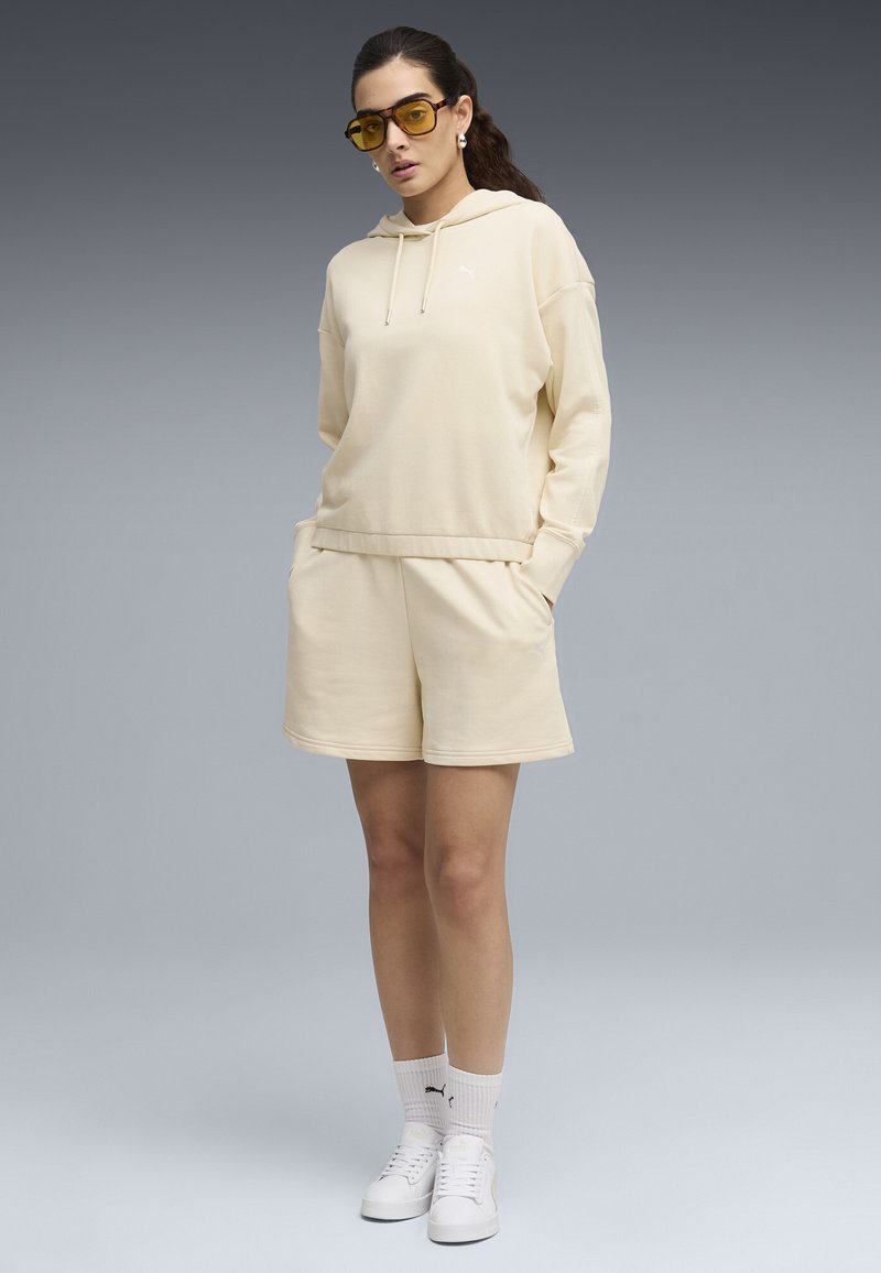 Femeie în picioare, purtând hanorac bej cu glugă și pantaloni scurți, adidași albi, șosete albe și ochelari de soare cu lentile galbene, pe fundal gri.