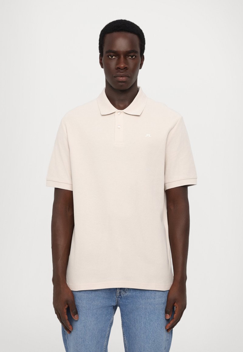 Beige polo-skjorte med myk tekstur, korte ermer og en tre-knappers placket. Har en liten hvit logo på brystet.