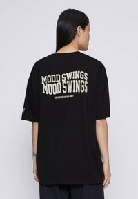 Camiseta negra de gran tamaño con un gran texto blanco impreso en la parte posterior que dice "CAMBIOS DE HUMOR" y un texto más pequeño debajo. Mangas cortas.