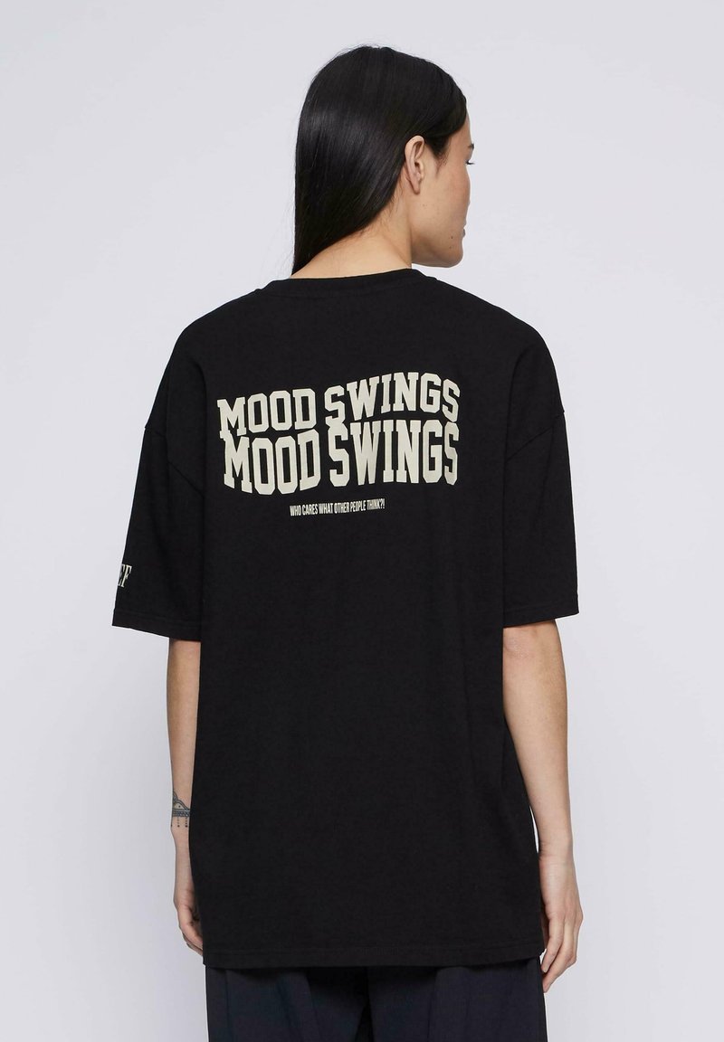 Camiseta negra de gran tamaño con un gran texto blanco impreso en la parte posterior que dice "CAMBIOS DE HUMOR" y un texto más pequeño debajo. Mangas cortas.
