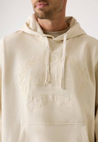 Beige kapučjaka ar izšūtu zīmola logo, dekoratīvu lauru un tekstu "KLASISKA AMERIKĀŅU STILS" uz krūtīm. Ieguvums ir priekšējā kabata.
