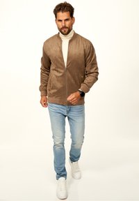 Braune Wildleder-Bomberjacke mit Reißverschluss, gerippten Bündchen, getragen über einem cremefarbenen Rollkragenpullover und hellblauen Jeans, kombiniert mit weißen Sneakers.