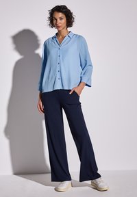 Camicia azzurra chiaro con maniche lunghe e colletto, abbinata a pantaloni a gamba larga blu scuro e sneakers bianche. Tessuto morbido, vestibilità rilassata.