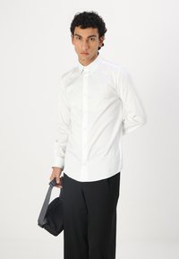 Camisa blanca de botones con cuello puntiagudo, combinada con pantalones de pierna ancha negros. El modelo sostiene un pequeño bolso negro con una correa gris.