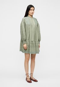 Γυναίκα με μακριά σκούρα μαλλιά φοράει ένα φαρδύ, ανοιχτό πράσινο φόρεμα με μακριά μανίκια και δαντελένιες λεπτομέρειες, καθώς και καφέ δερμάτινα παπούτσια με κορδόνια, στέκεται σε λευκό φόντο.