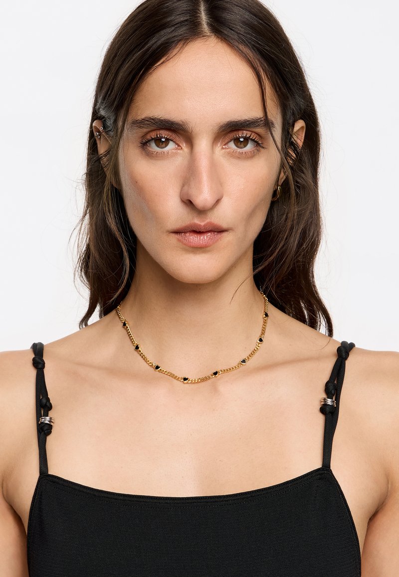 Collar de cadena dorada con acentos de piedras negras, presentando un diseño delicado. La camiseta sin mangas negra con tirantes anudados complementa la joyería.