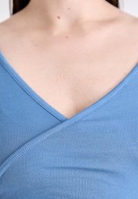 Ribani, svijetloplavi top s V-izrezom i preklopom na prsima, s glatkom teksturom i finim detaljima šivanja.