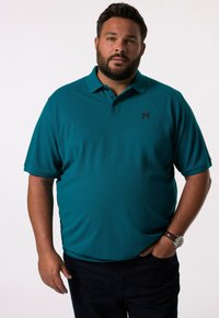 Turkusowa koszulka polo z krótkimi rękawami, kołnierzykiem i czarnym wyszywanym logo. Model nosi ciemne dżinsy i zegarek na nadgarstku. Gładkie białe tło.