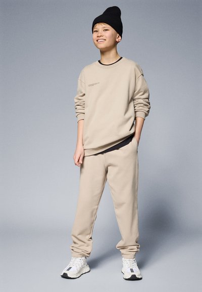 Sudadera beige y joggers a juego, hechos de una tela suave, con una camiseta negra debajo. Presenta un dobladillo acanalado y un gorro de lana negro.