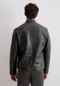 Mann mit lockigem, dunklem Haar, der eine schwarze Lederjacke und dunkelgraue Hosen trägt und vor einer schlichten weißen Wand steht.