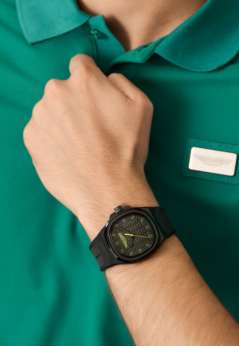 Montre bracelet noire avec un cadran noir texturé, des accents verts et un bracelet en caoutchouc, portée sur une main près d'un polo vert avec un emblème blanc.