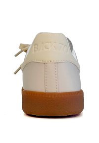 Sneaker bianca con una parte superiore in tela liscia, una suola in gomma testurizzata e il testo in rilievo "BACK70" sul tallone. Palette di colori neutri.