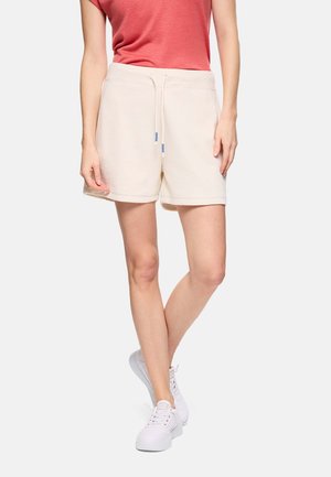 Mujer con shorts beige con cordón, camiseta coral de manga corta y zapatillas blancas, de pie con las piernas cruzadas sobre un fondo liso.