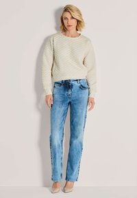 Vrouw met kort blond haar, gekleed in een gequilt crème trui, lichte blauwe rechte jeans en beige puntige hakken, staande tegen een eenvoudige muur.