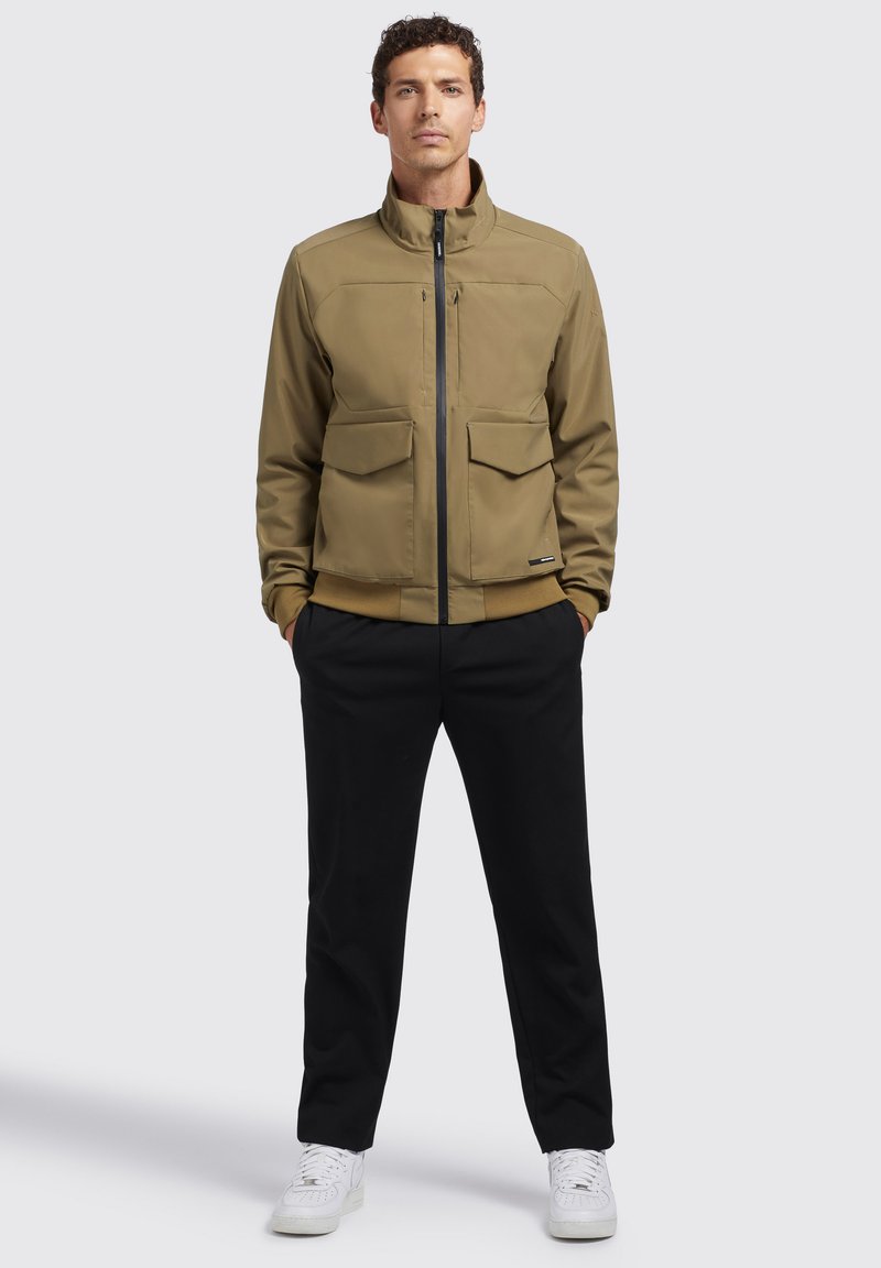 Veste vert olive clair avec un col montant, deux poches avant et une fermeture éclair. Un pantalon noir et des baskets blanches complètent la tenue.