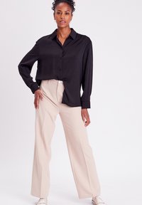 Chemise noire à manches longues et à boutons, avec une coupe décontractée, associée à un pantalon large beige clair et des baskets beiges sur un fond uni.