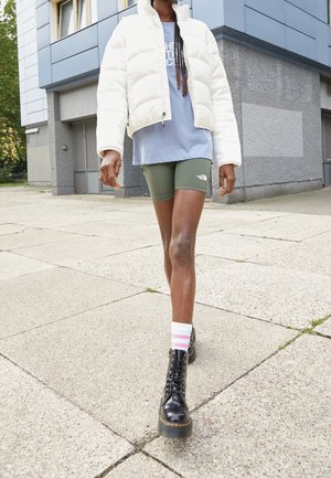 Personne marchant sur le trottoir portant une doudoune blanche, un t-shirt bleu, un short vert, des chaussettes rayées et des bottes noires à lacets devant un bâtiment moderne.