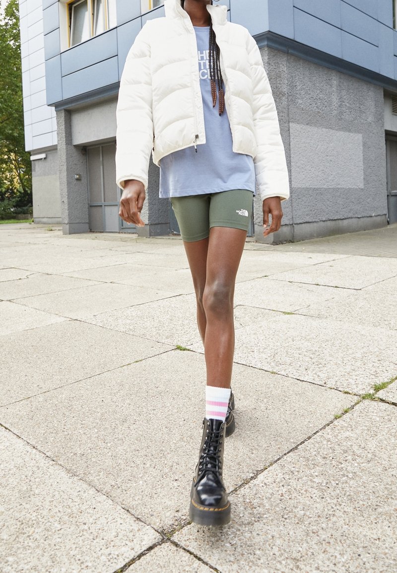 Personne marchant sur le trottoir portant une doudoune blanche, un t-shirt bleu, un short vert, des chaussettes rayées et des bottes noires à lacets devant un bâtiment moderne.