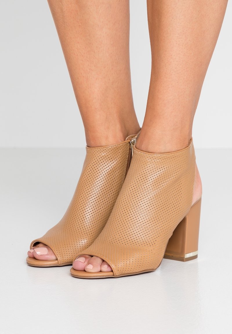 Femme portant des bottines beige perforées à bout ouvert avec talons carrés et fermetures éclair latérales sur un fond de sol gris clair.
