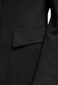 Schwarzer, maßgeschneiderter Blazer mit glatter Textur, einer aufgesetzten Tasche am Ärmel und einem klaren Nahtdesign. Nahaufnahme hebt die Details hervor.