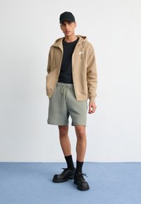Sweatshirt bege com fecho, t-shirt preta, calções verde-claro, meias pretas e sapatilhas pretas robustas. Design simples com bolsos frontais e cordão ajustável.