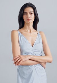 Rochie fără mâneci, de culoare albastră deschisă, cu un material texturat, decolteu în V și detalii pliate în față, asortată cu o bijuterie simplă.