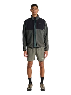 Młody mężczyzna stoi, ubrany w szarą i czarną kurtkę polarową na zamek błyskawiczny, zieloną koszulkę, oliwkowe szorty z nadrukiem "ATW", czarne skarpetki i beżowe sneakersy.