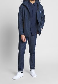Giacca impermeabile blu navy con cappuccio, chiusura a zip e tasche laterali, indossata sopra una camicia blu navy con bottoni, abbinata a jeans di denim scuro e sneakers bianche.
