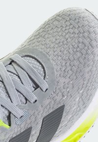 Gros plan d'une chaussure de sport grise en tissu texturé, avec des lacets gris assortis et des accents jaunes sur une semelle blanche.