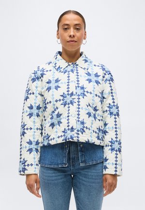 Frau trägt eine gesteppte weiße Jacke mit blauen Sternmustern über Jeans, steht vor einem schlichten hellen Hintergrund.