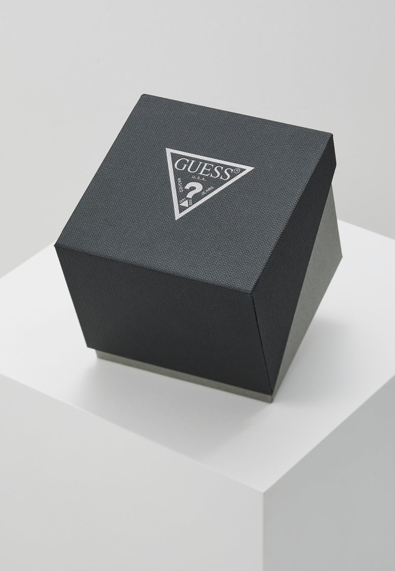Caja de regalo texturizada negra con forma triangular, que presenta un logo plateado de "GUESS" y un signo de interrogación en la parte superior, situada sobre un pedestal blanco.